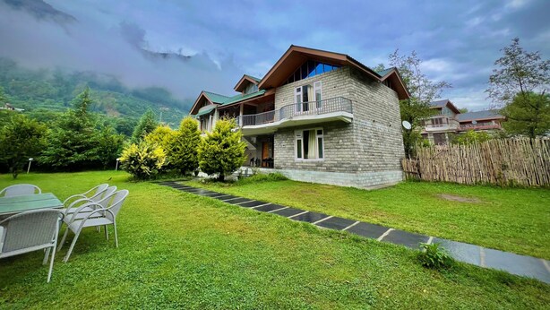 A two-story stone house with a balcony, surrounded by a well-maintained lawn and a wooden fence. - Raison Meadows (Kullu)