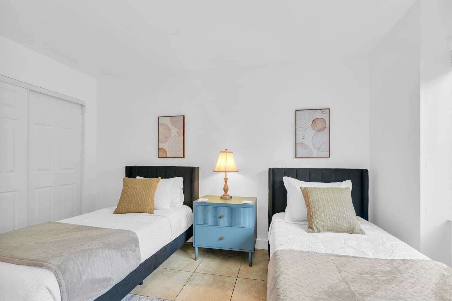 A hotel room with two single beds, a nightstand with a lamp, and two framed artworks on the walls.