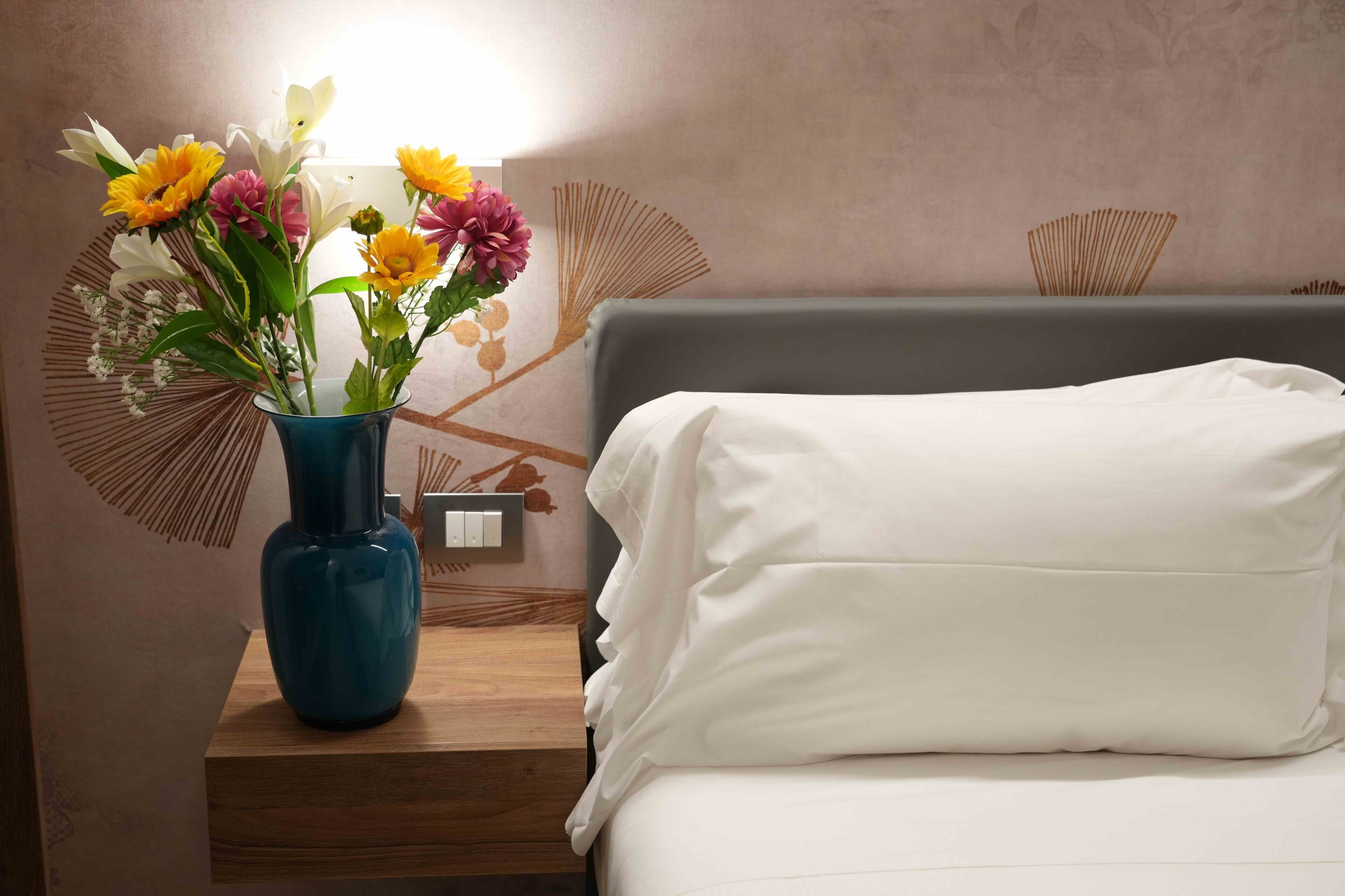 Una cama con ropa blanca, un jarrón azul con flores y una mesita de noche de madera.