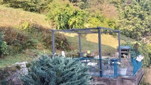 Un gazebo con struttura in metallo e tetto, circondato da vegetazione e una collina sullo sfondo.