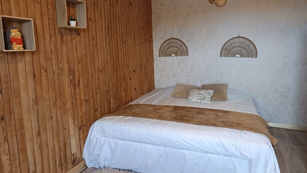 Una camera da letto con una parete in legno, un letto con biancheria bianca, due applique decorative e un piccolo ripiano con una pianta.