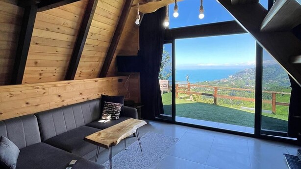 Ein modernes Loft-Zimmer mit schrägem Dach, Holzbalken, einem grauen Sofa, einem Holztisch und einem großen Fenster mit Blick auf eine grüne Landschaft.