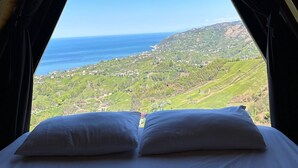 Ein Bett mit weißer Bettwäsche und Kissen, in einem Raum mit einem dreieckigen Fenster, das Blick auf eine Küstenlandschaft bietet.