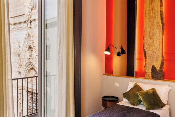Habitación de hotel con cama, almohadas, mesita de noche, lámpara y un amplio ventanal con vistas a un edificio histórico.