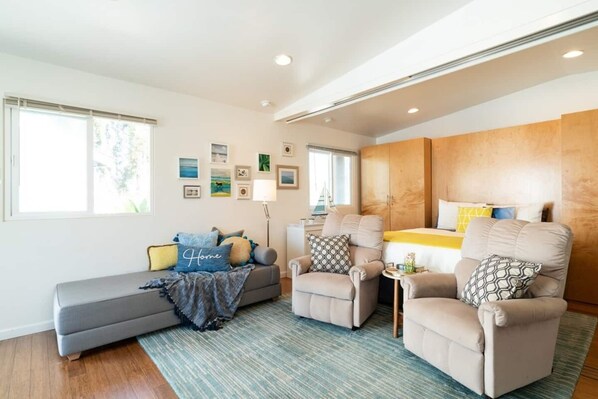 A modern living room with a sofa, two armchairs, a small table, and a bed with a wooden headboard. There are framed pictures on the wall and a window providing natural light.