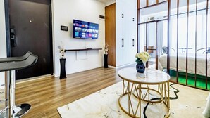 A hotel room with a wooden floor, a round table with a vase of flowers, a TV mounted on the wall, and a sliding door leading to a balcony. - Alba Garden (Nairobi)