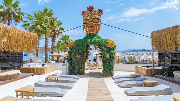 Un espace de détente aménagé sur la plage, avec des parasols en chaume, des chaises longues et une grande arche décorative.