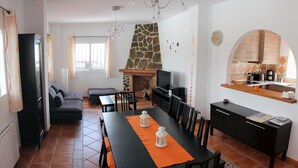 A modern living room with a fireplace, a dining area with a long table, and a kitchen with a stone backsplash. - El Escalador - Detached finca in the mountains outside Competa (Cómpeta)