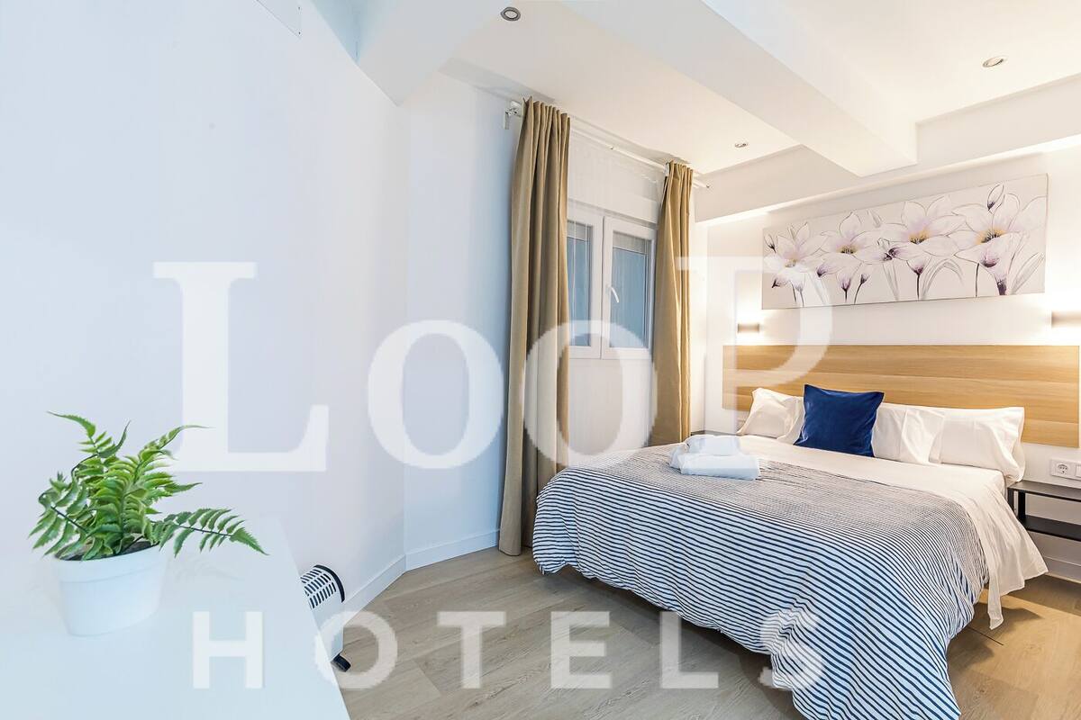 A hotel room with a bed, curtains, a potted plant, and a wall with a floral painting.