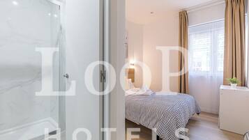 Quarto de hotel com cabine de chuveiro de vidro, cama com roupa de cama listrada, mesa de cabeceira com uma planta e janela com cortinas.