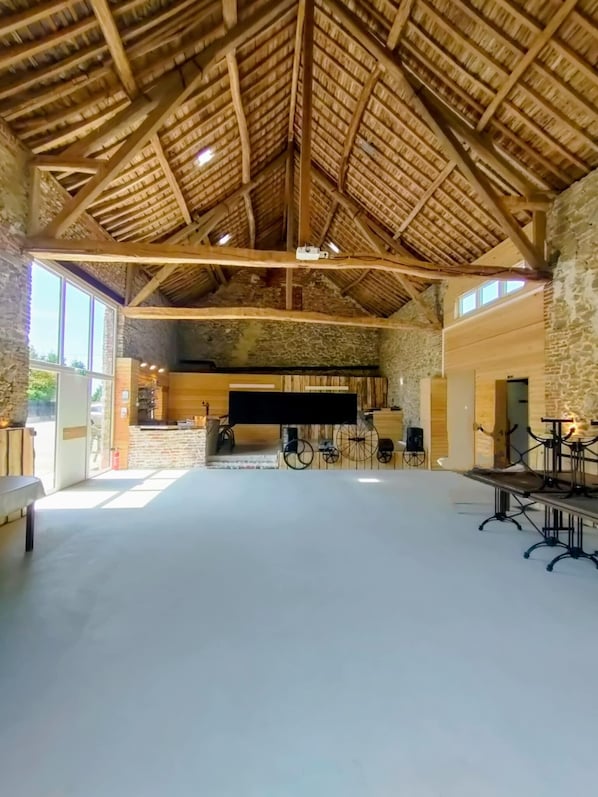 A spacious hall with a high wooden ceiling and stone walls. There is a long table with chairs, a large screen, and a bicycle wheel decoration.