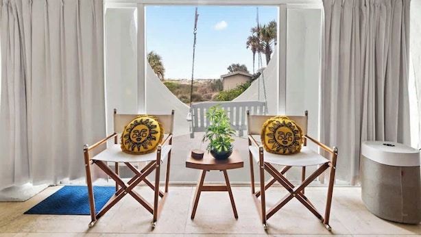 A room with a view through a large window, two chairs with sun-themed cushions, a small table with a plant, and a trash bin.