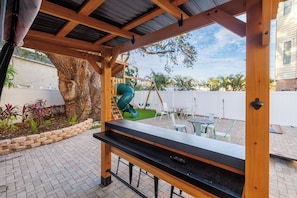 Relax in a shaded patio area with a pergola, table, and chairs, surrounded by a natural tree and a children's play area nearby.