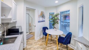 A compact kitchen with a sink, microwave, and stove, a dining area with a round table and blue chairs, and a window with blinds.