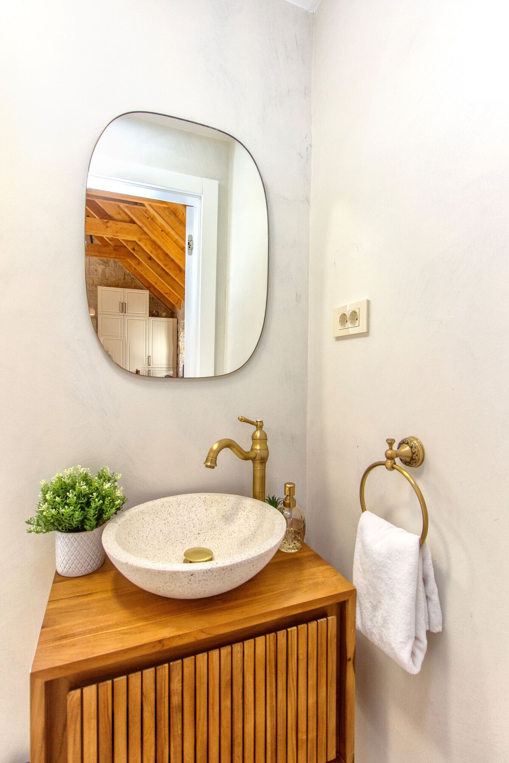 A bathroom with a round, speckled sink, a wooden vanity, a gold faucet, a round mirror, a towel ring, and a small potted plant.