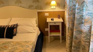 Una habitación de hotel con una cama, una mesita de noche con lámpara, una pequeña mesa y una cortina con motivos naturales.