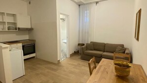 Ein kompakter Wohnbereich mit Küchenzeile, einem Essbereich mit Holztisch und -stühlen sowie einem Wohnzimmer mit Sofa und Lampe.