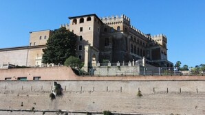 A large, historic building with a prominent tower and arched windows, situated next to a fortified wall with sparse vegetation.