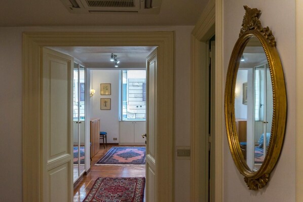 A hallway with a large ornate mirror, wooden flooring, and a view into a room with a bed and a window.