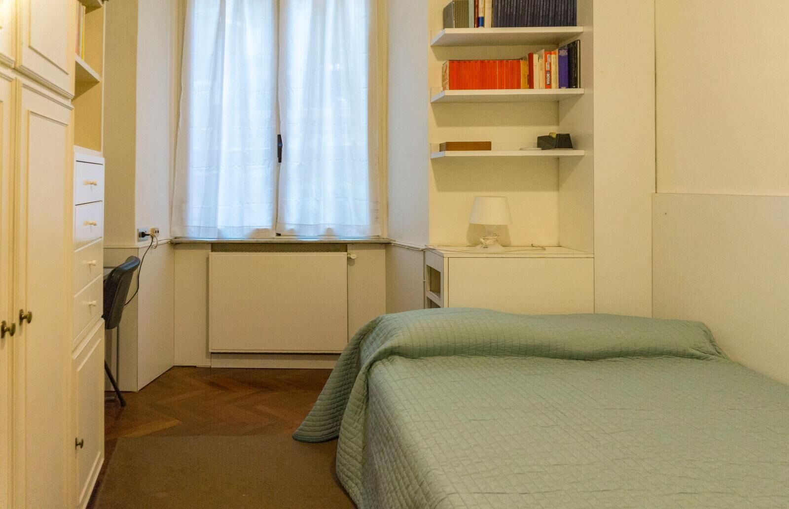 Een compacte, keurig opgeruimde slaapkamer met een bed, een radiator, een boekenplank en een raam met gordijnen.
