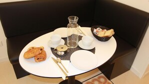 A small round table with a white tablecloth, a black cushioned bench, a glass pitcher, two white cups, a small metal teapot, a bowl of bread, and a plate with croissants.