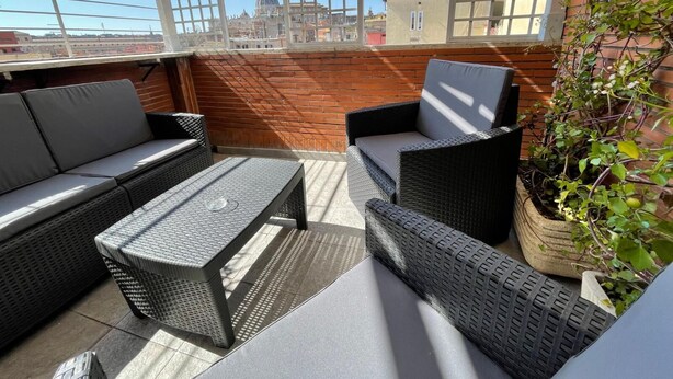 Un patio al aire libre con mobiliario de mimbre, una planta en maceta y vistas de un paisaje urbano.