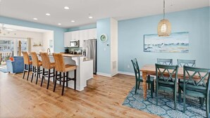 A modern kitchen with white cabinets, a central island with bar stools, and a dining area with a wooden table and chairs. - Harborview Retreat Mansion (Ocean City)