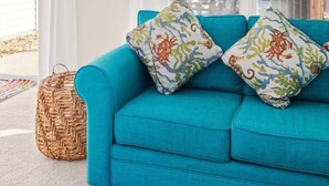 A bright living room with a teal sofa, two patterned cushions, a woven basket, and a view of houses through large windows. - Harborview Retreat Mansion (Ocean City)