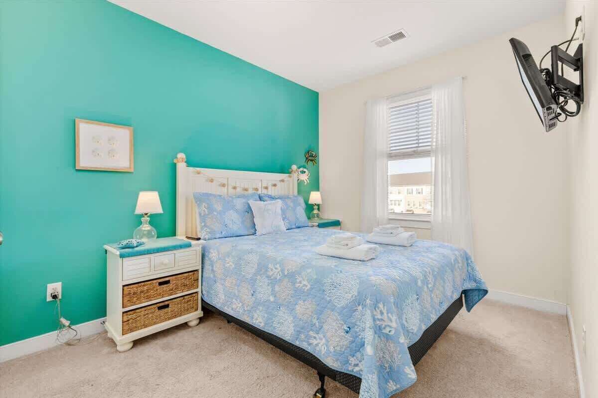 Una camera da letto con una parete color petrolio, un letto bianco con biancheria azzurra, un comodino con una lampada e una finestra con persiane.