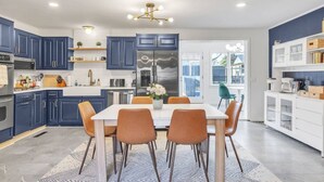 Une cuisine moderne avec des meubles bleus, un réfrigérateur en acier inoxydable et un coin repas comprenant une table blanche et des chaises orange.