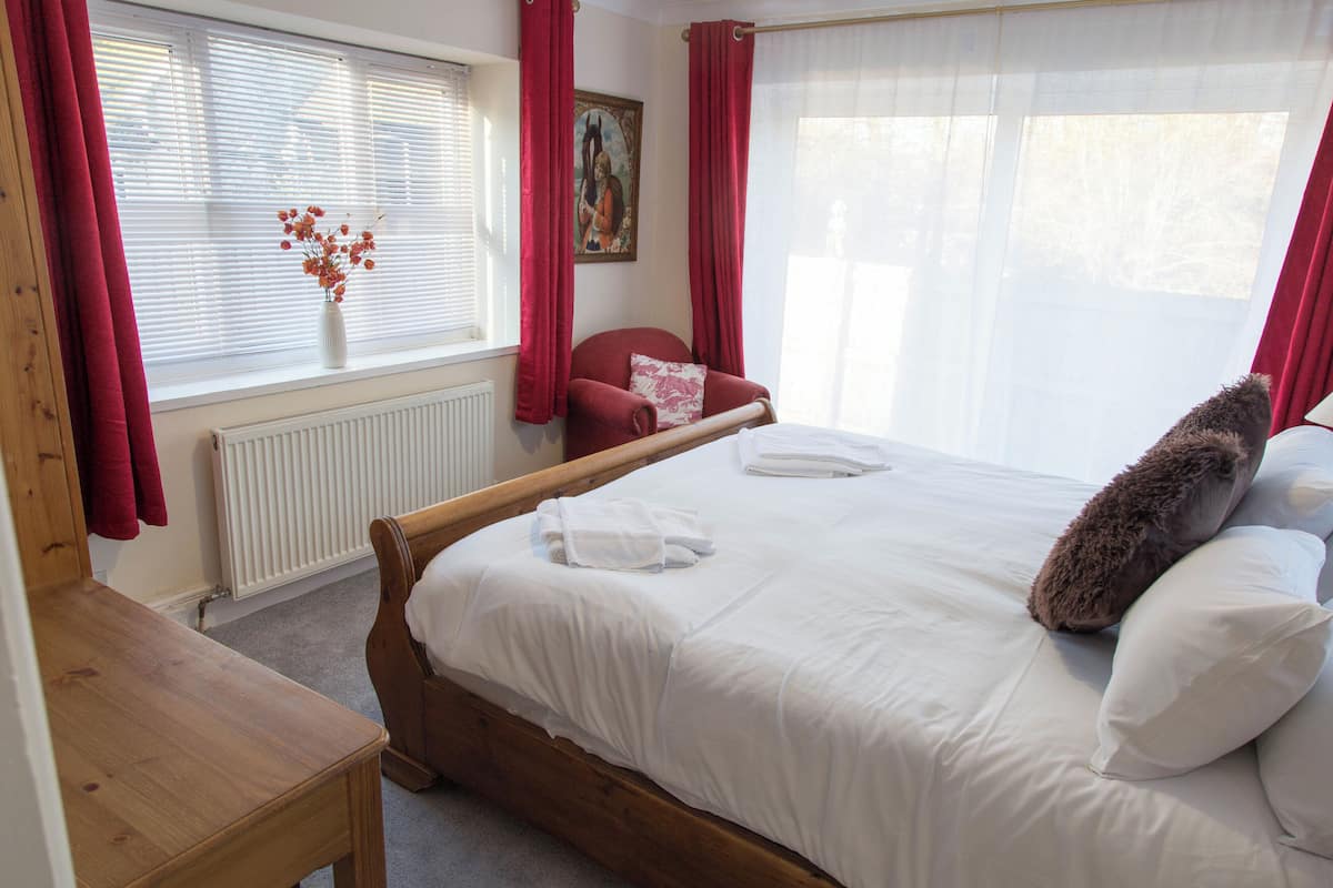 Een slaapkamer met een houten bed, witte beddengoed, rode gordijnen, een radiator en een schilderij aan de muur.