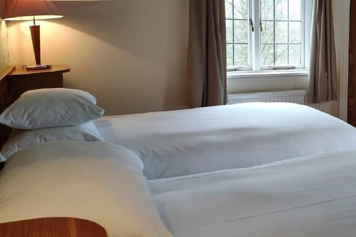 Een hotelkamer met twee bedden, een nachtkastje met een lamp, een raam met gordijnen en een houten deur.