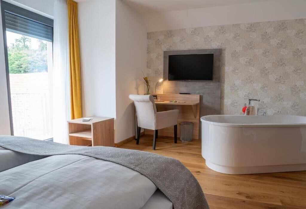 A modern hotel room with a bed, a desk with a chair, a TV, and a bathtub.