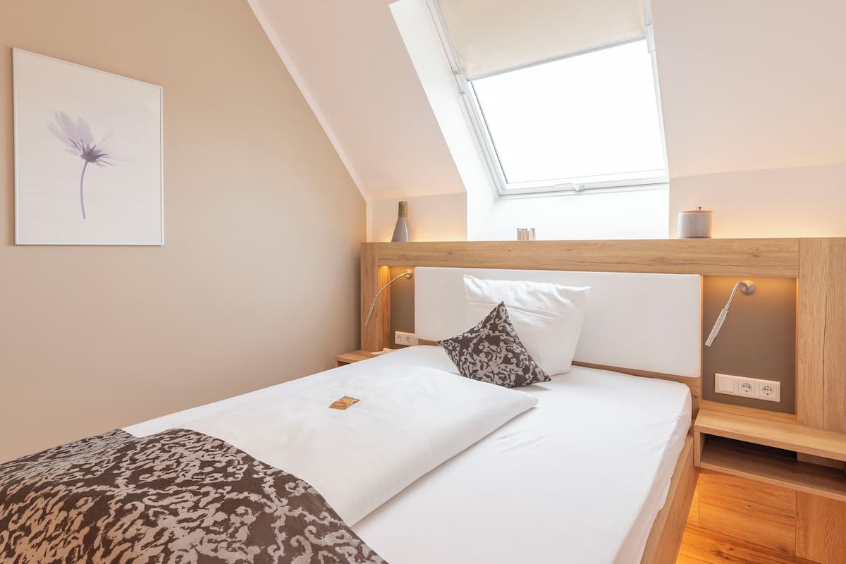 A bedroom with a sloped ceiling, a skylight, a bed with white bedding, a patterned pillow, and a framed artwork on the wall.