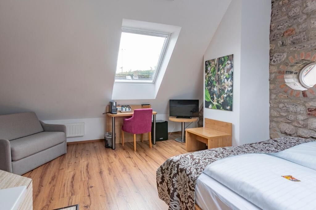 A modern room with a sloped ceiling, a bed, a desk with a computer, a chair, and a television.
