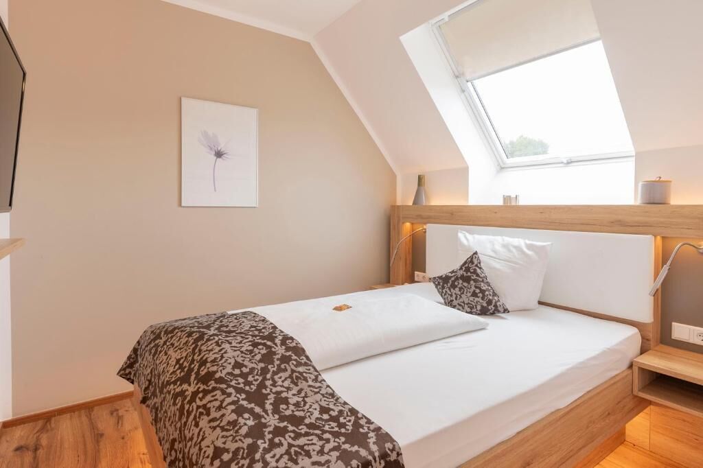 Ein Schlafzimmer mit Schräge, einem Oberlicht, einem Bett mit gemusterten Textilien, einem Nachttisch und einem Wandbild.
