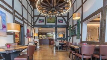 A restaurant with a high ceiling, exposed beams, and a central chandelier. Tables and chairs are arranged for dining.