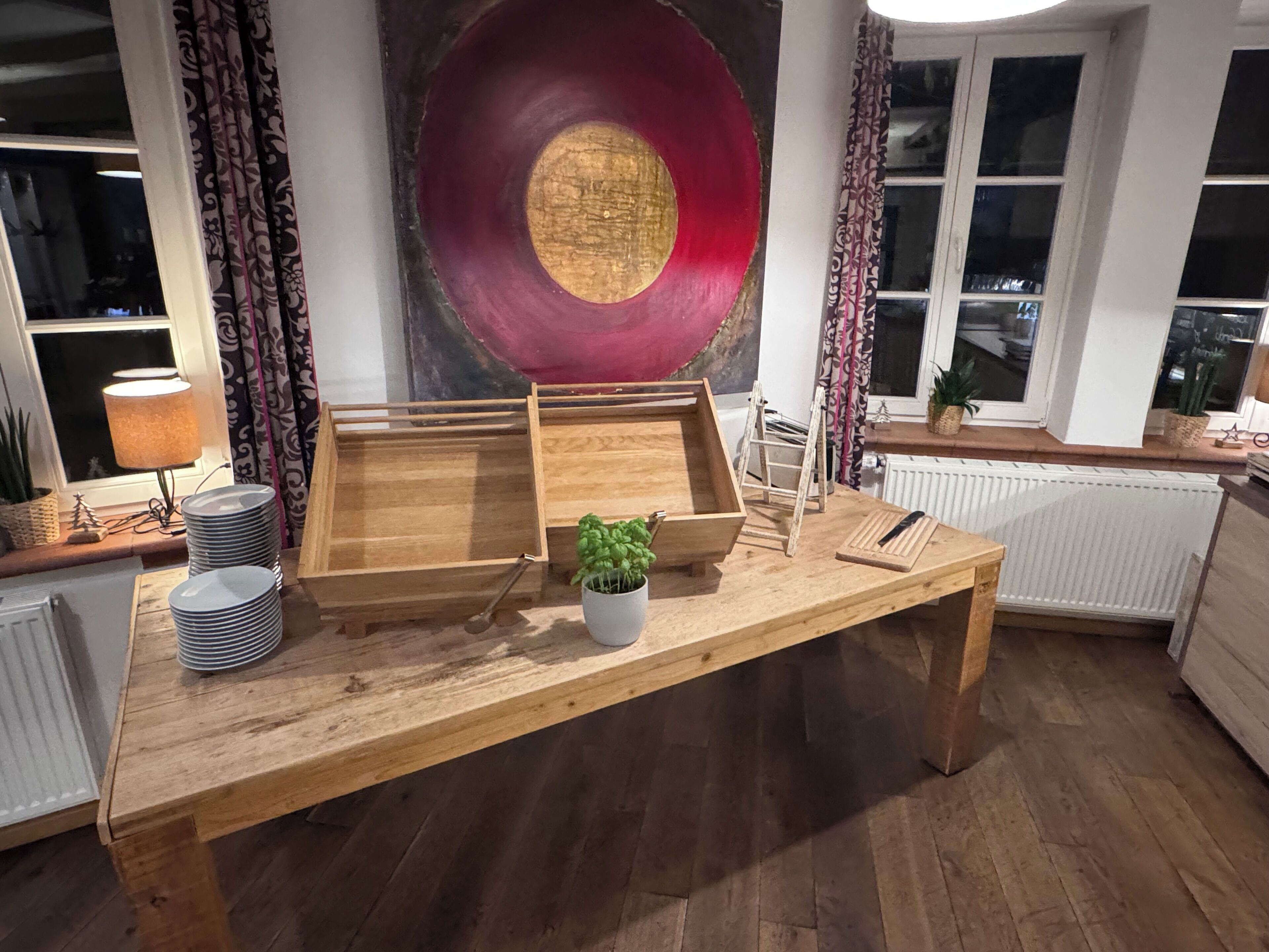 Una mesa de madera con bandejas, una planta en maceta y una lámpara en una habitación con cortinas estampadas y un cuadro circular grande en la pared.