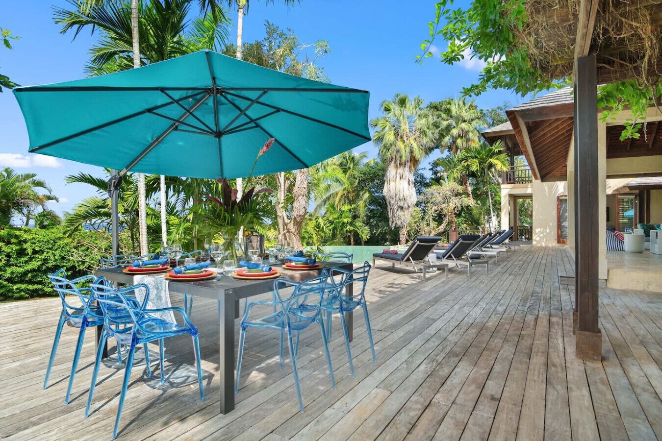 Um deck de madeira com uma mesa posta para refeições, rodeado por cadeiras azuis e um grande guarda-sol verde oferecendo sombra.