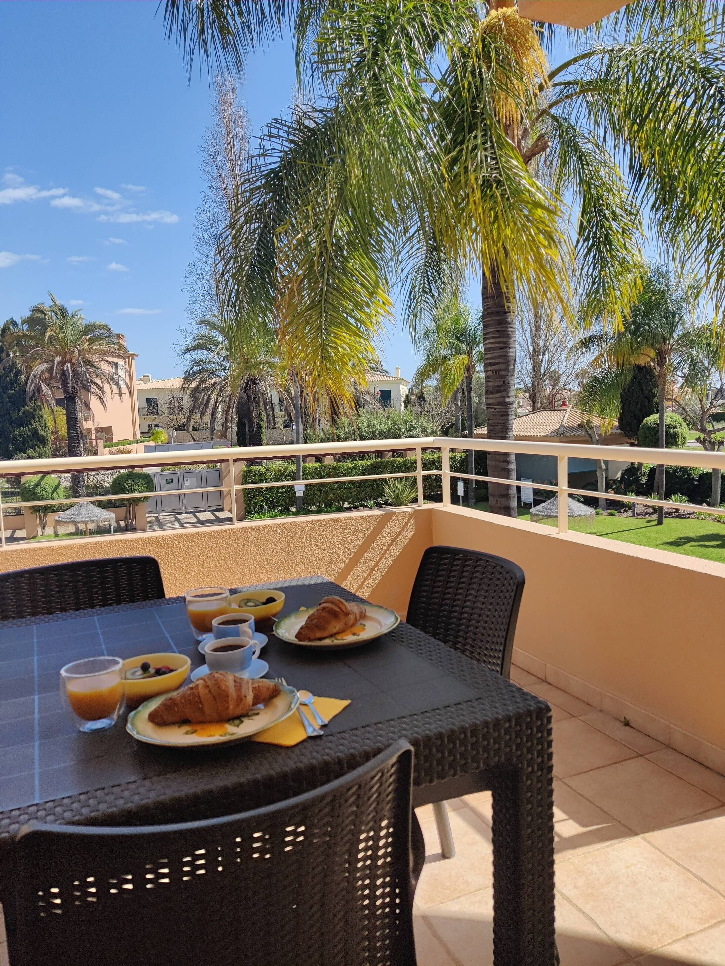 A balcony with a table set for breakfast, overlooking a garden with palm trees and a building in the background.