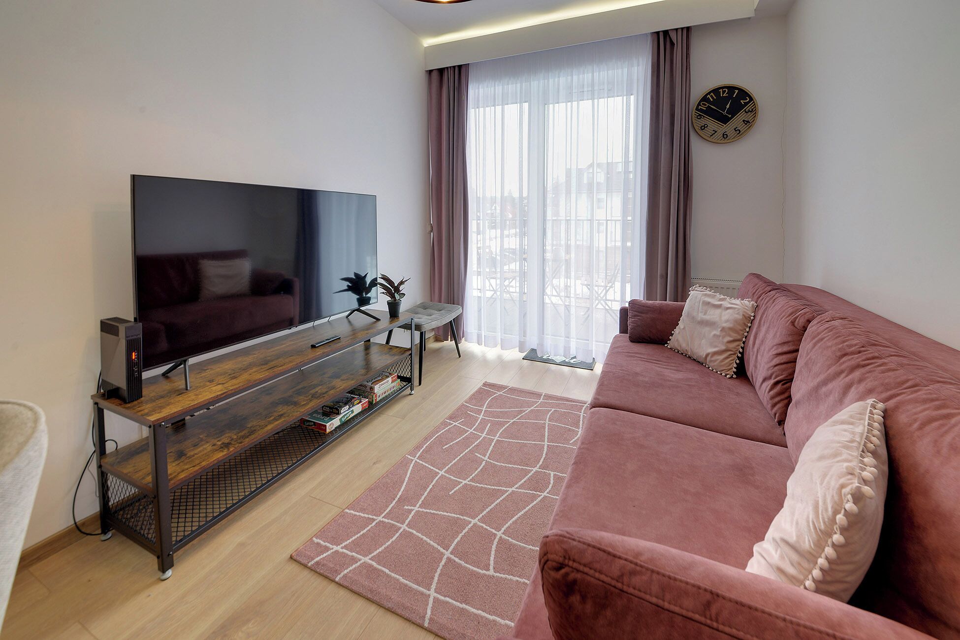 A modern living room with a flat-screen TV, a wooden entertainment unit, a pink sofa, and a large window with sheer curtains.