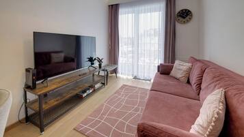 A modern living room with a flat-screen TV, a wooden entertainment unit, a pink sofa, and a large window with sheer curtains.