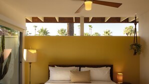 Une chambre d’hôtel avec un lit, un ventilateur de plafond, une fenêtre donnant sur des palmiers et une plante suspendue.
