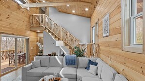 A cozy cabin interior with a wooden staircase, a gray sectional sofa, and a dining area with outdoor seating. - Bear Paw Getaway - Beech luxury with hot tub, fire pit (Beech Mountain)