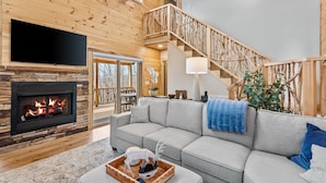 A cozy living room with a stone fireplace, a grey sofa, a wooden staircase, and a large TV. - Bear Paw Getaway - Beech luxury with hot tub, fire pit (Beech Mountain)