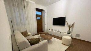 A modern living room with a sofa, coffee table, and ottoman, a flat-screen TV mounted on the wall, and a small side table with decorative items. - Cozy Corner (Nova Milanese)