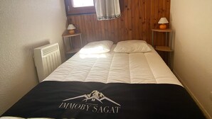 Un dormitorio con cama, ventana con cortinas, radiador y lámpara en una mesita de noche.