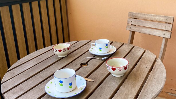 Una mesa de madera con cuatro tazas y platillos blancos, cada uno decorado con motivos florales en rojo y azul, en un balcón con barandilla de madera y vistas al follaje.