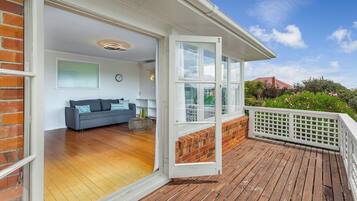 An open sliding door leading to a living room with a sofa and a round coffee table, a wooden deck, and a view of greenery and a red-roofed building.