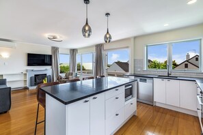 A modern kitchen with a central island, white cabinetry, and a large window offering a view of the neighborhood. - Renovated Home with Lovely Garden by Zodiak Stay (Auckland)